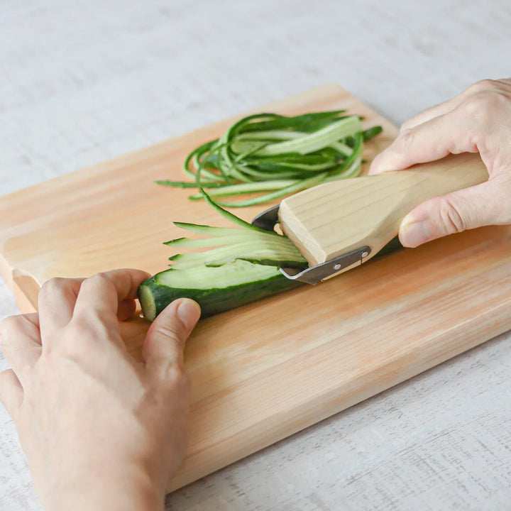 Hands slicing a cucumber into thin julienne strips using a Japanese wood handle julienne peeler on a board.
