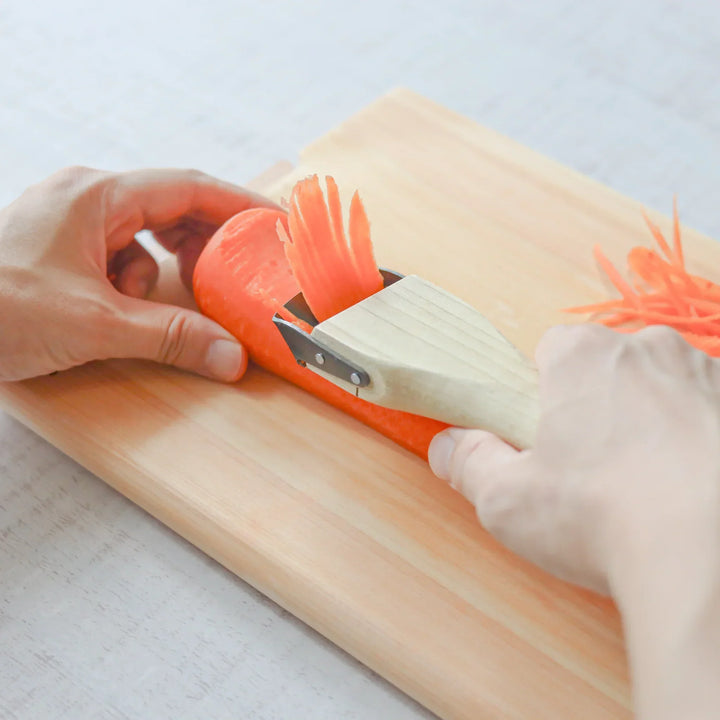 Hands creating julienne carrot strips with a Japanese wood handle julienne peeler on a wooden cutting board.
