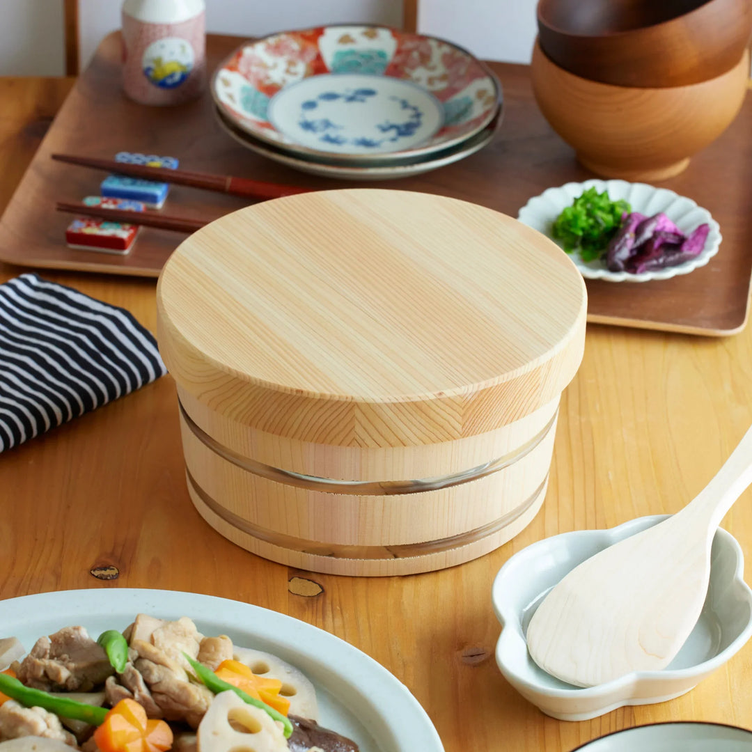Lidded wooden ohitsu rice storage bowl placed on a Japanese dining table with chopsticks, side dishes, and a rice paddle.